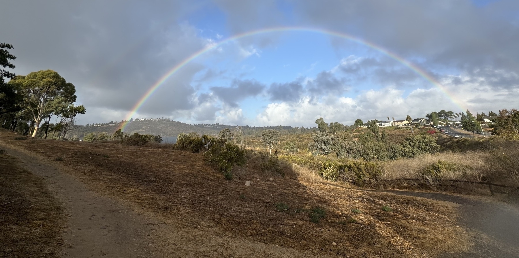 A rainbow arches over an overlook of a lake, with shrubs and grass in the foreground and a path arcing away and downward, under a partly cloudy sky.