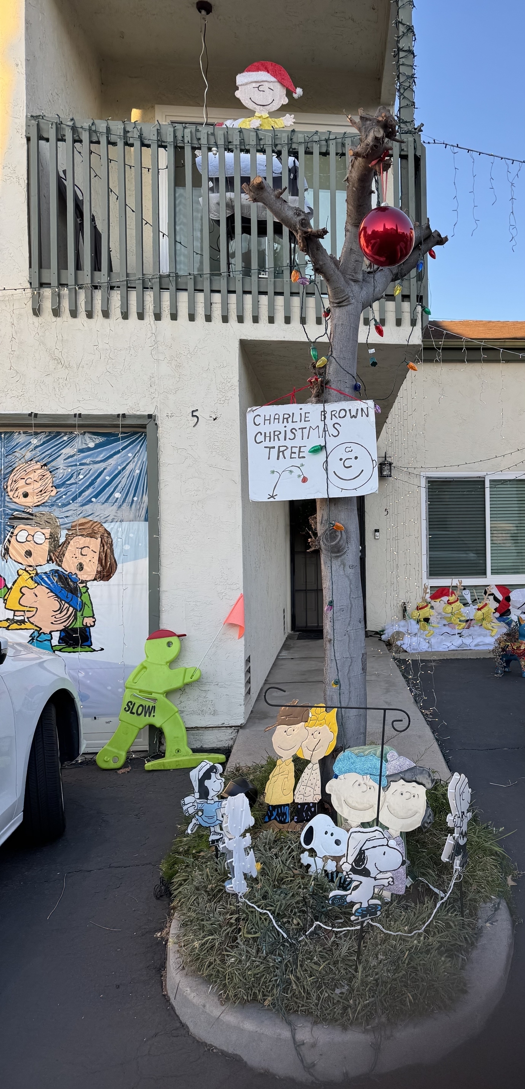 A bare tree about 10 feet tall, on a grassy island in a parking lot in front of a condo, with a couple of ornaments on top and Charlie Brown figures at the base. A sign on the tree reads, ‘Charlie Brown's Christmas Tree.’