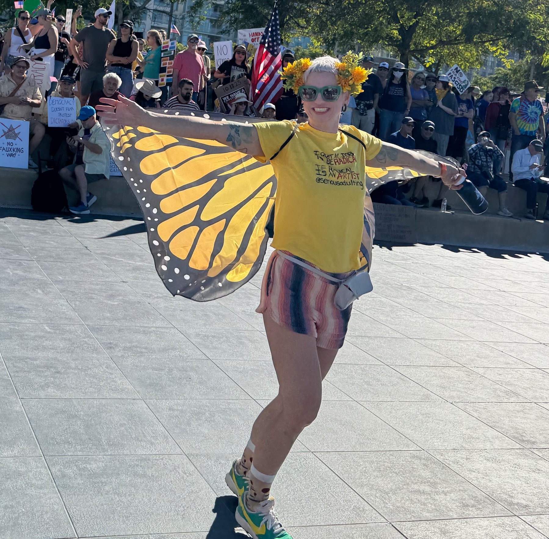 Auto-generated description: A person dressed in a yellow butterfly costume with flowers in their hair is smiling and posing in an outdoor gathering with a crowd holding signs in the background.
