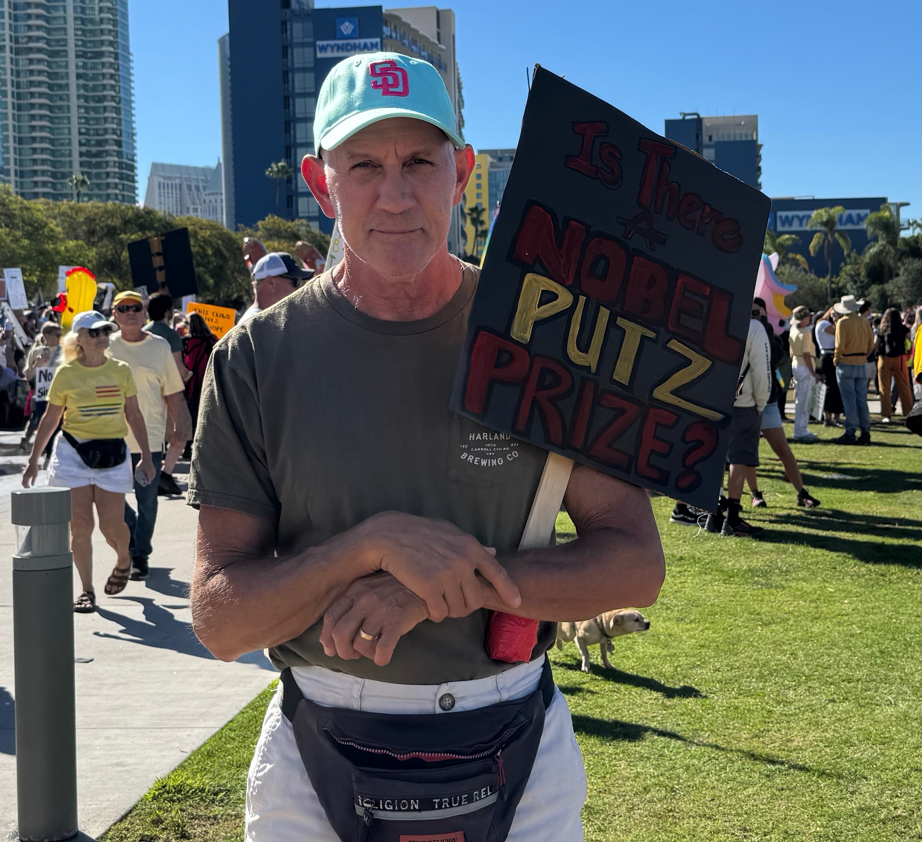Auto-generated description: A person is holding a sign that reads Is There a Nobel Putz Prize? while standing in a park during a protest or gathering.