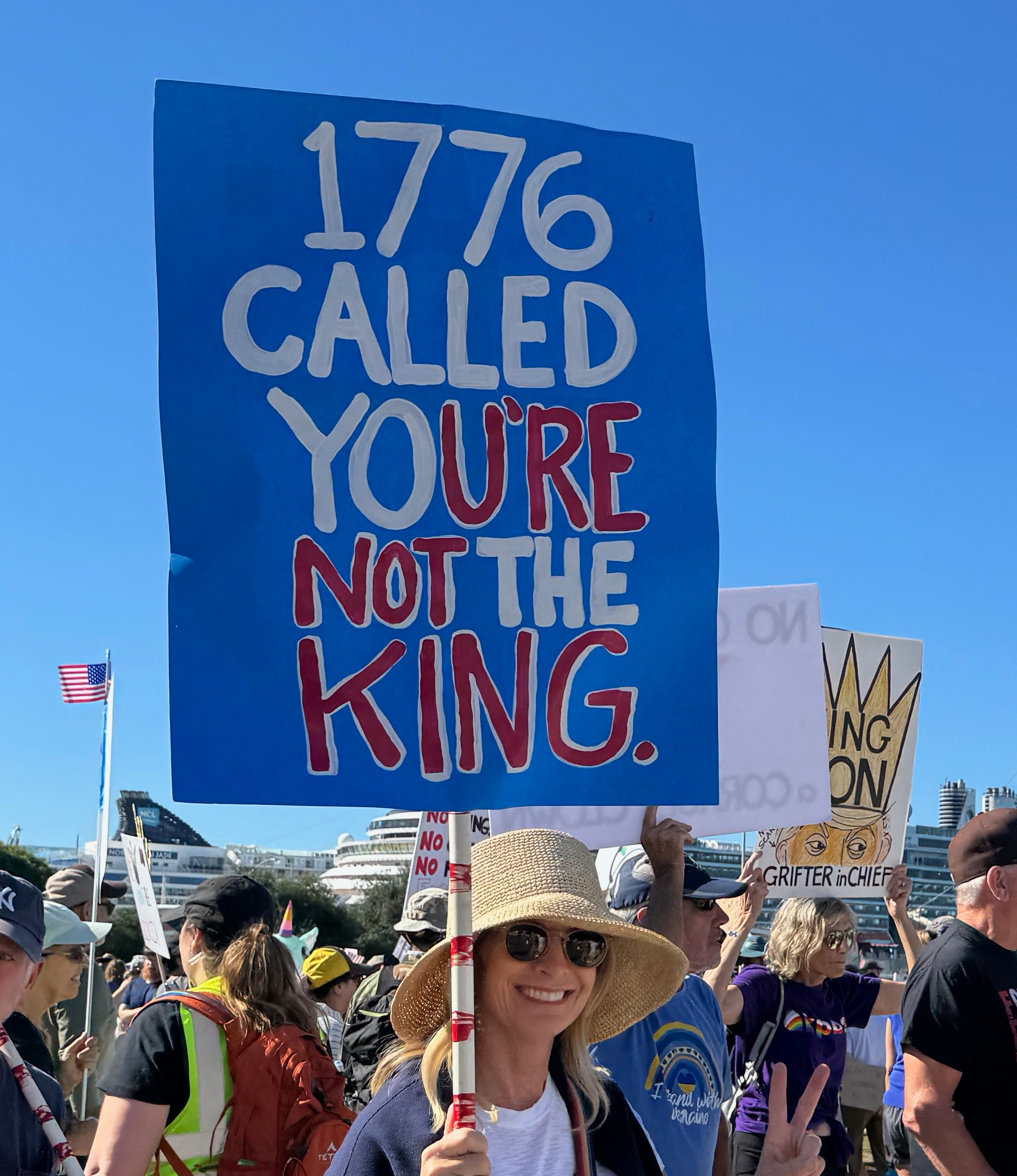 Auto-generated description: A woman wearing a straw hat and sunglasses holds a sign that reads 1776 CALLED YOU’RE NOT THE KING amidst a crowd and flags.