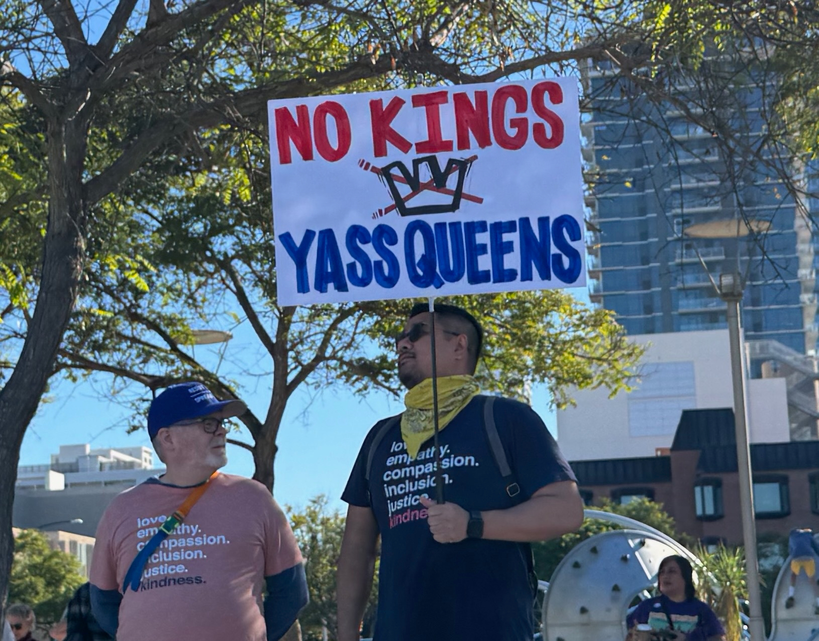 Auto-generated description: Two individuals stand outdoors holding a sign that reads NO KINGS YASS QUEENS, with buildings and trees in the background.