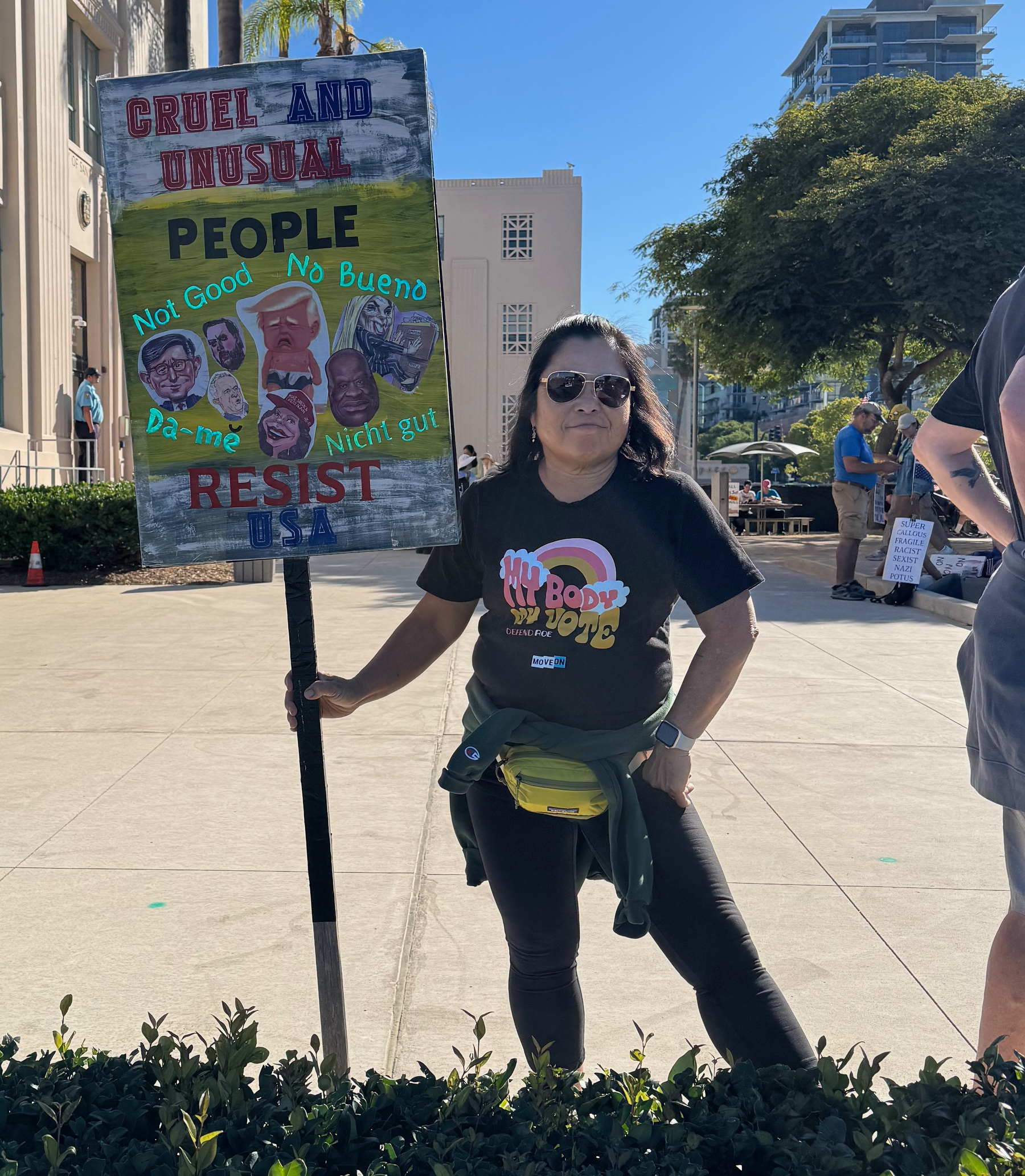 Auto-generated description: A person stands outdoors holding a sign that reads Cruel and Unusual People with cut-out heads and phrases like No Good and No Bueno, wearing a My Body, My Choice T-shirt.