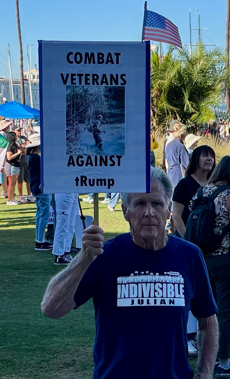 Auto-generated description: A man is holding a sign that reads Combat Veterans Against Trump at an outdoor gathering.