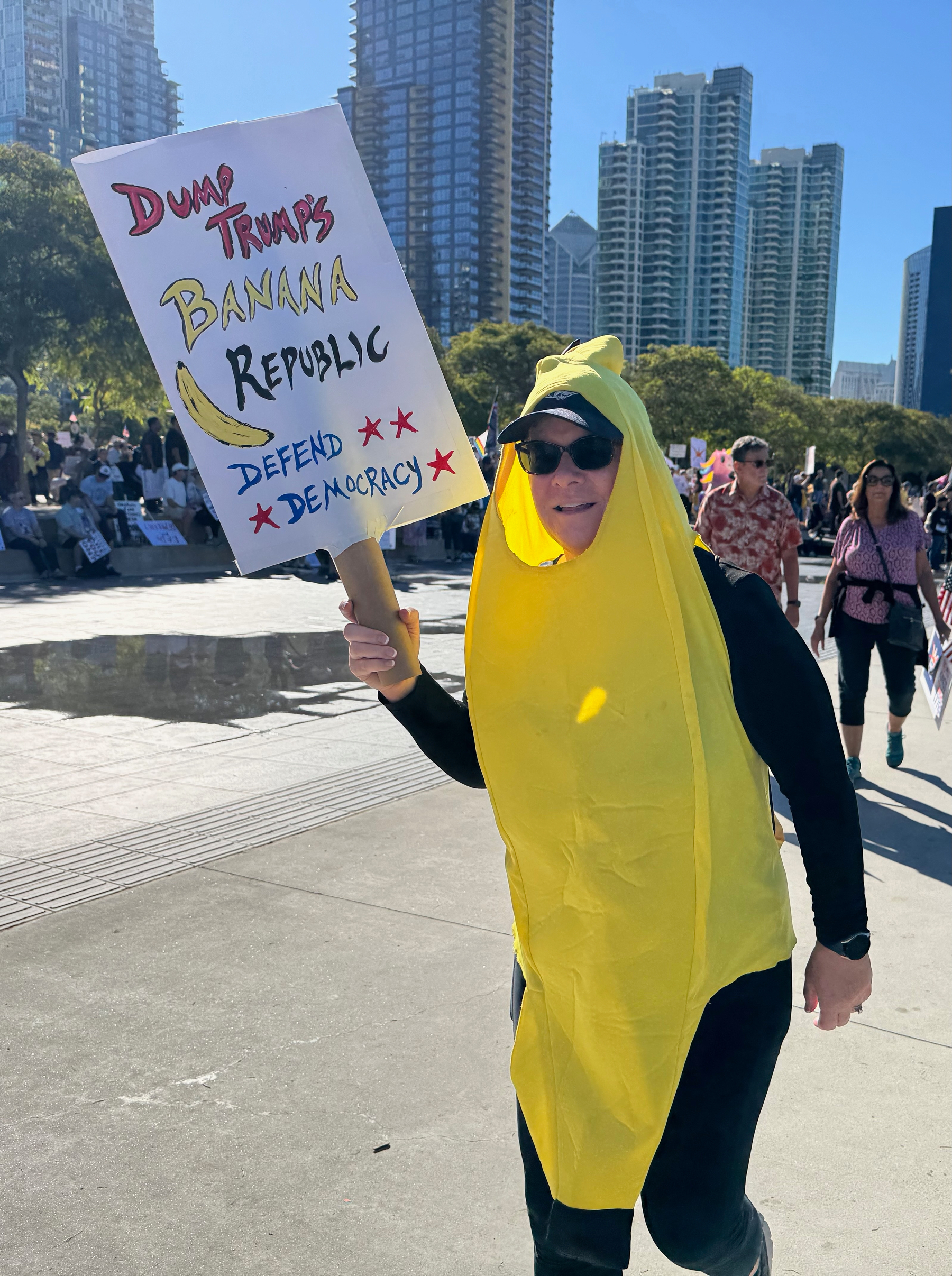 Auto-generated description: A person dressed in a banana costume holds a protest sign that reads, Dump Trump's Banana Republic, while walking among a crowd in a cityscape setting.