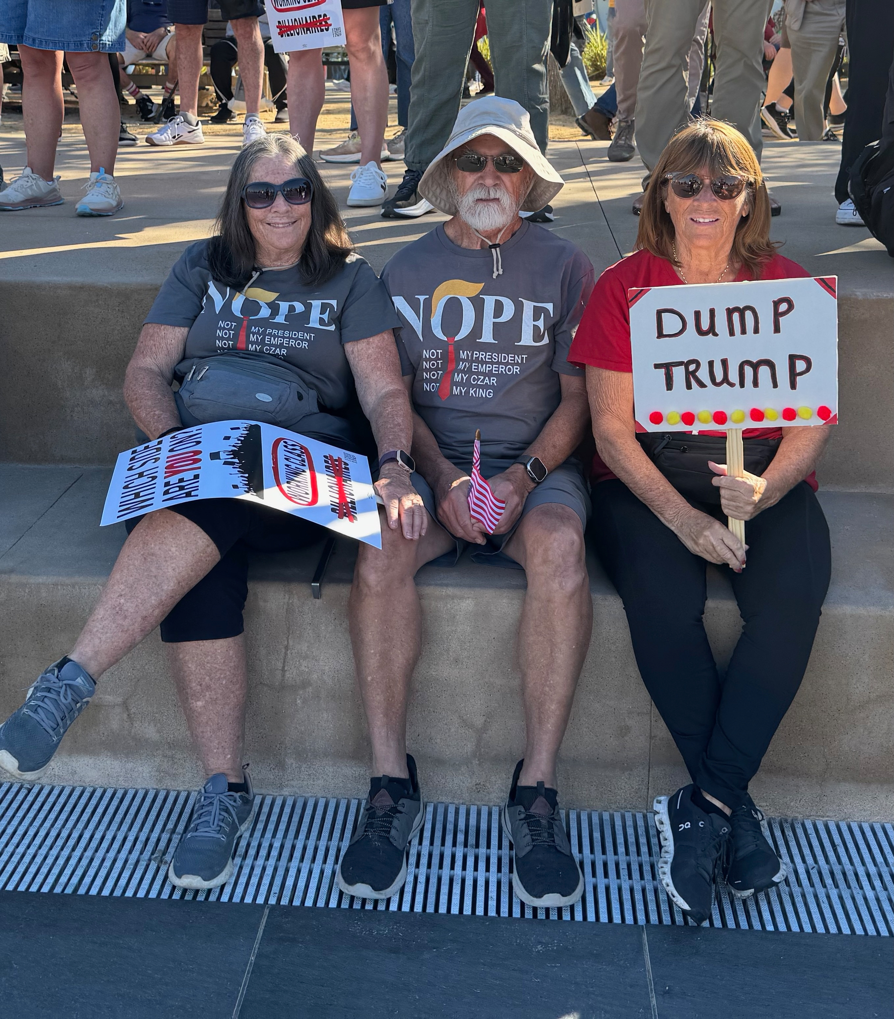 Auto-generated description: Three people are sitting on a ledge holding signs, with two wearing shirts that say NOPE and the third holding a sign that says Dump Trump.