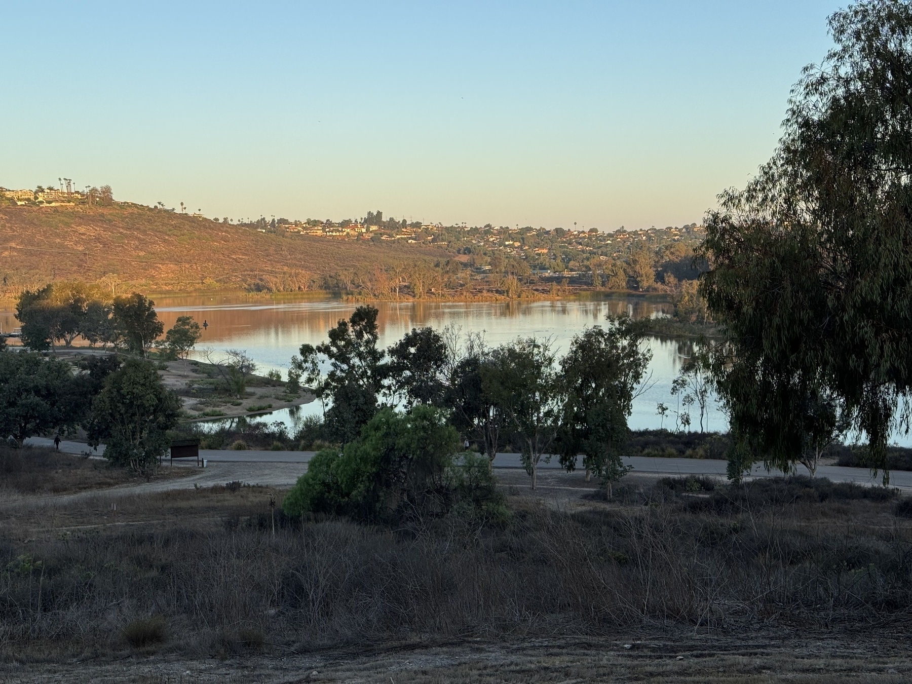 A peaceful lake at dawn, surrounded by trees and hills under a clear sky, with scattered buildings visible in the distance.